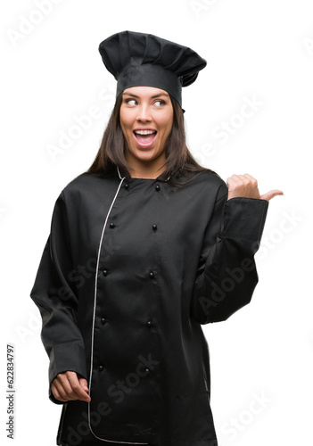 Young hispanic cook woman wearing chef uniform pointing and showing with thumb up to the side with happy face smiling