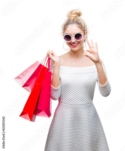 Young beautiful blonde woman holding shopping bags over isolated background doing ok sign with fingers, excellent symbol