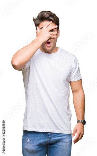 Young handsome man wearing white t-shirt over isolated background peeking in shock covering face and eyes with hand  looking through fingers with embarrassed expression.