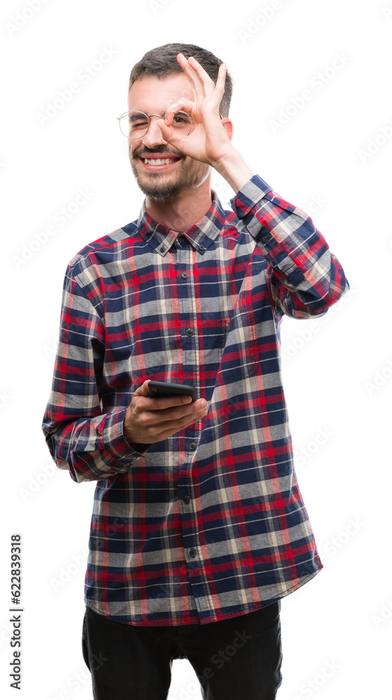 Young hipster adult man holding smartphone with happy face smiling doing ok sign with hand on eye looking through fingers Young hipster adult man holding smartphone with happy face smiling doing ok sign with hand on eye looking through fingers