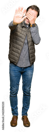 Middle age handsome man wearing winter vest covering eyes with hands and doing stop gesture with sad and fear expression. Embarrassed and negative concept.