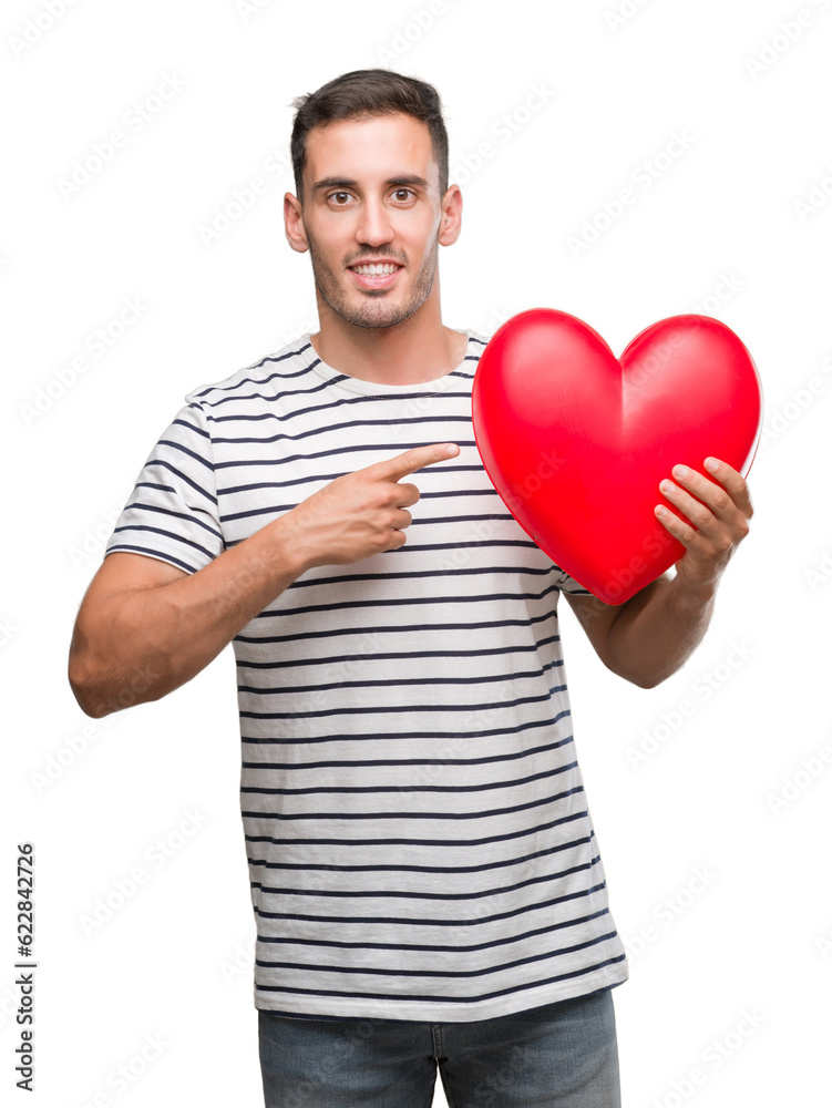 Handsome young man in love holding red heart very happy pointing with hand and finger Handsome young man in love holding red heart very happy pointing with hand and finger