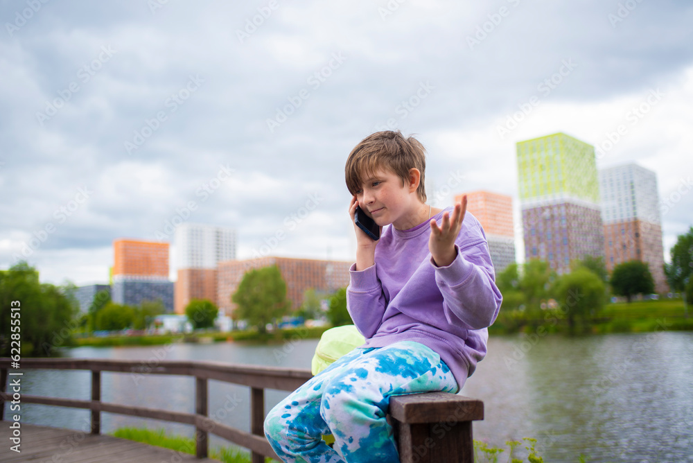 emotional boy talking using a smartphone, calling his parents or friends, a schoolboy is sitting on a fence on the river embankment in the city and talking on a mobile phone.