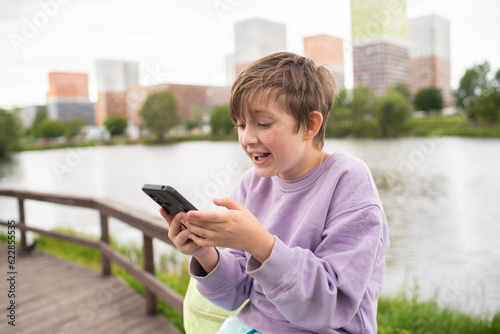 emotional child plays games on the phone, communicates with friends online, watches streams. boy sits on the embankment of the river in the city, after school.