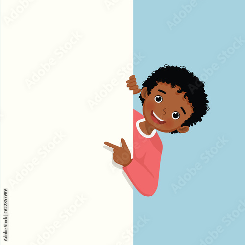 Little African boy standing behind blank board peeking and pointing to empty banner with finger
