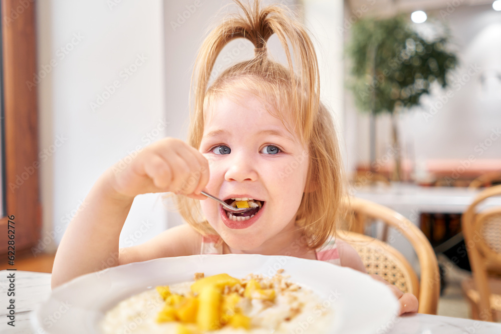 Little kid having breakfast at cafe. Adorable girl drinking still water, eating rice porridge with mango. enjoying breakfast. Little kid having breakfast at cafe. Adorable girl drinking still water, eating rice porridge with mango. enjoying breakfast.