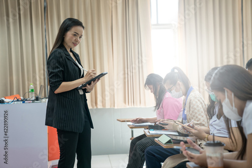 A university lecturer wearing a business suit holds a tablet while teaching and explaining lessons to students in a classroom