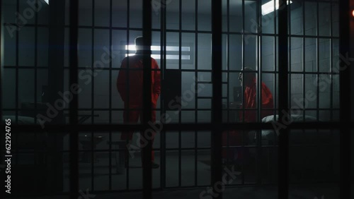 Two guilty inmates in detention center or correctional facility. Elderly criminal in orange uniform sits on jail bed and talks with cellmate. Gloomy prisoners serve imprisonment term in prison cell.