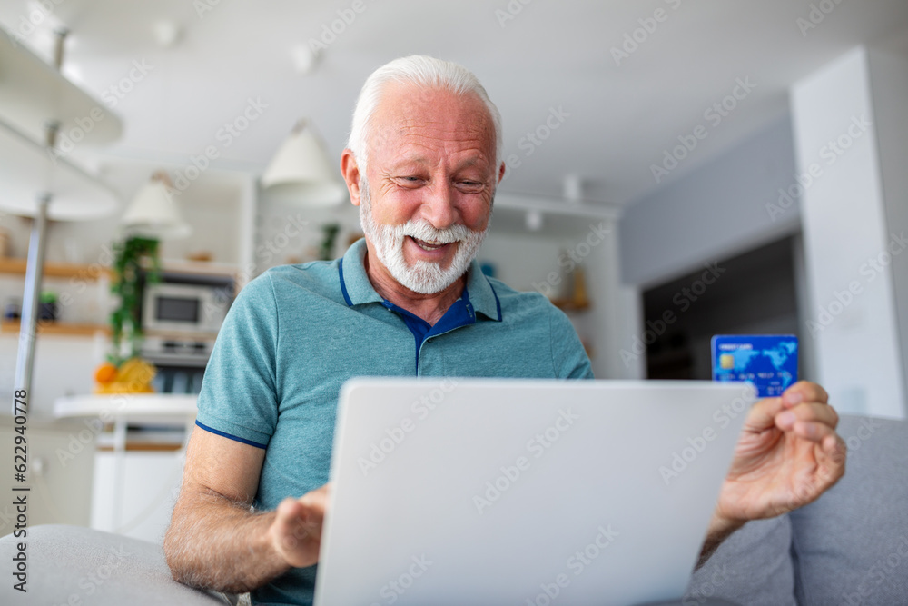 Mature man using laptop, holding plastic credit or debit card, senior grey haired customer making secure internet payment, shopping or browsing online banking service, entering information Mature man using laptop, holding plastic credit or debit card, senior grey haired customer making secure internet payment, shopping or browsing online banking service, entering information