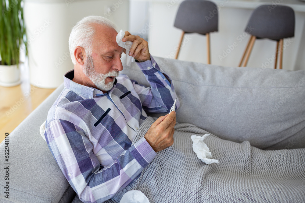 Sick elderly man checking his temperature suffering from seasonal flu or cold lying on sofa caughing suffering from seasonal flu or cold. . Ill senior feel unhealthy with influenza at home Sick elderly man checking his temperature suffering from seasonal flu or cold lying on sofa caughing suffering from seasonal flu or cold. . Ill senior feel unhealthy with influenza at home