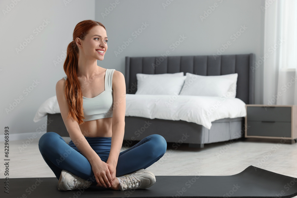 Young woman in sportswear sitting on fitness mat in bedroom, space for text