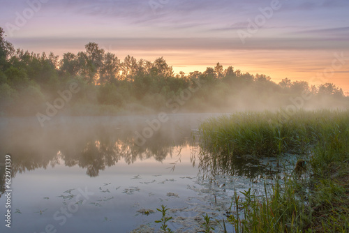 Foggy haze creeps over the river on a summer morning at dawn.