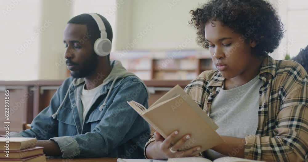 African American people students sitting at desk in library woman reading book and man listening to music and using laptop typing. Campus and youth lifestyle concept.