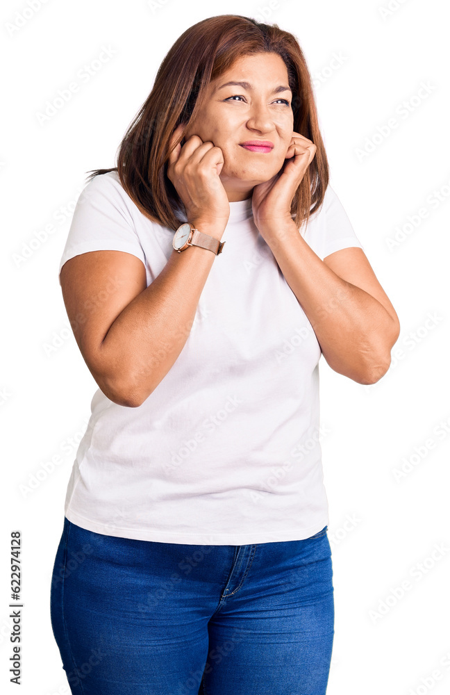 Middle age latin woman wearing casual white tshirt covering ears with fingers with annoyed expression for the noise of loud music. deaf concept. Middle age latin woman wearing casual white tshirt covering ears with fingers with annoyed expression for the noise of loud music. deaf concept.