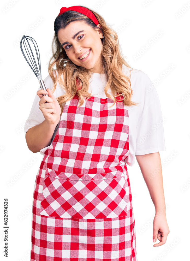 Young caucasian woman wearing apron holding whisk looking positive and happy standing and smiling with a confident smile showing teeth Young caucasian woman wearing apron holding whisk looking positive and happy standing and smiling with a confident smile showing teeth