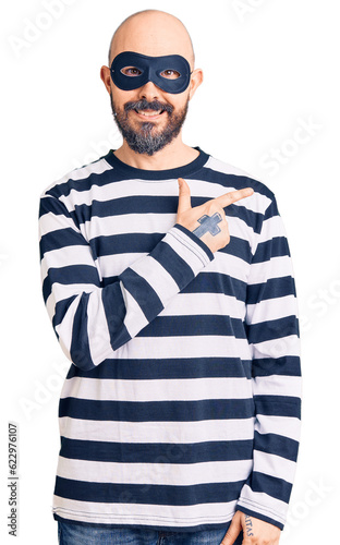 Young handsome man wearing burglar mask cheerful with a smile on face pointing with hand and finger up to the side with happy and natural expression © Krakenimages.com