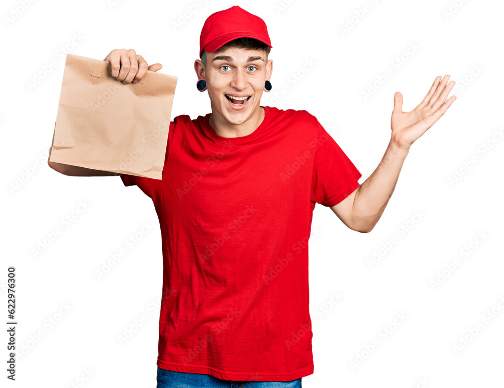 Young caucasian boy with ears dilation holding take away paper bag celebrating victory with happy smile and winner expression with raised hands