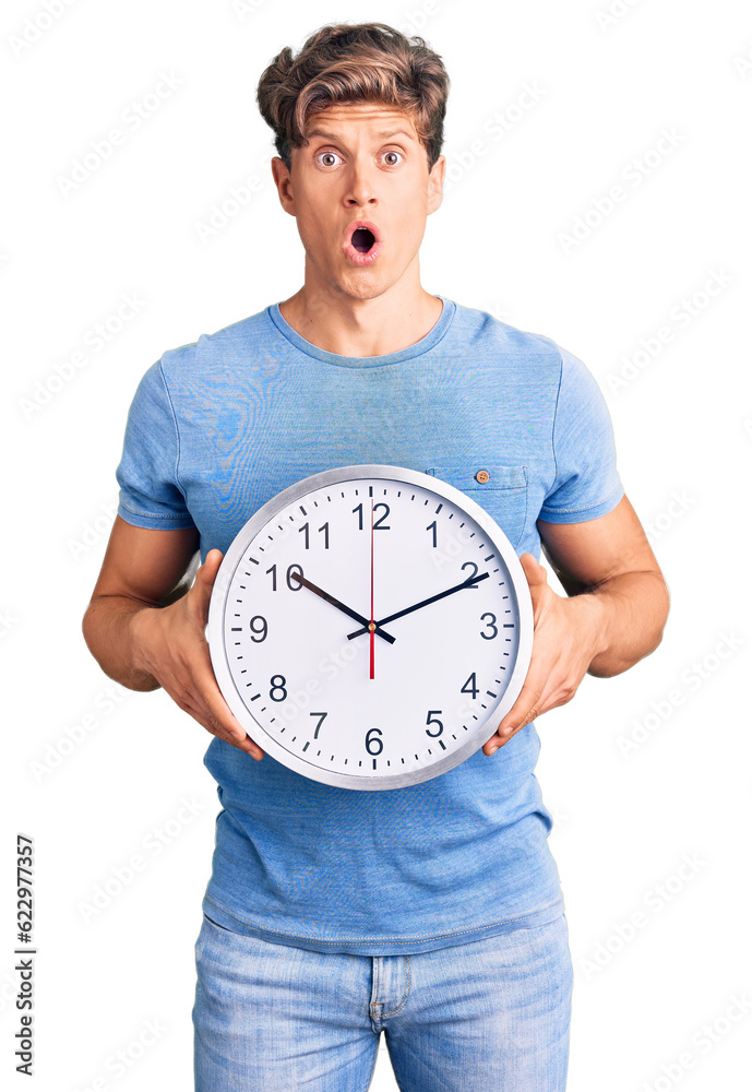 Young handsome man holding big clock scared and amazed with open mouth for surprise, disbelief face Young handsome man holding big clock scared and amazed with open mouth for surprise, disbelief face