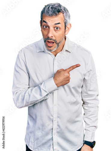 Middle age hispanic with grey hair wearing casual white shirt surprised pointing with finger to the side, open mouth amazed expression.