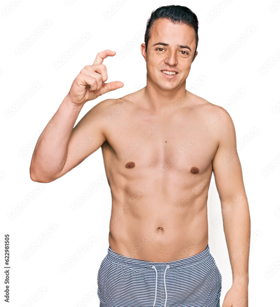 Handsome young man wearing swimwear shirtless smiling and confident gesturing with hand doing small size sign with fingers looking and the camera. measure concept. Handsome young man wearing swimwear shirtless smiling and confident gesturing with hand doing small size sign with fingers looking and the camera. measure concept.