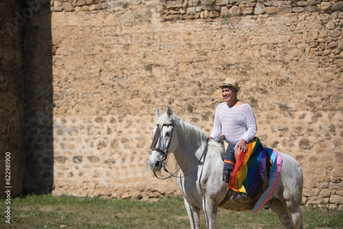 Non-binary person  young and South American  very makeup  mounted on a white horse  smiling and happy  with a gay pride flag on the rump  next to an old medieval castle. Concept queen  lgbtq   pride.