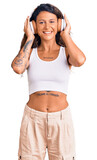Young hispanic woman with tattoo listening to music using headphones looking positive and happy standing and smiling with a confident smile showing teeth