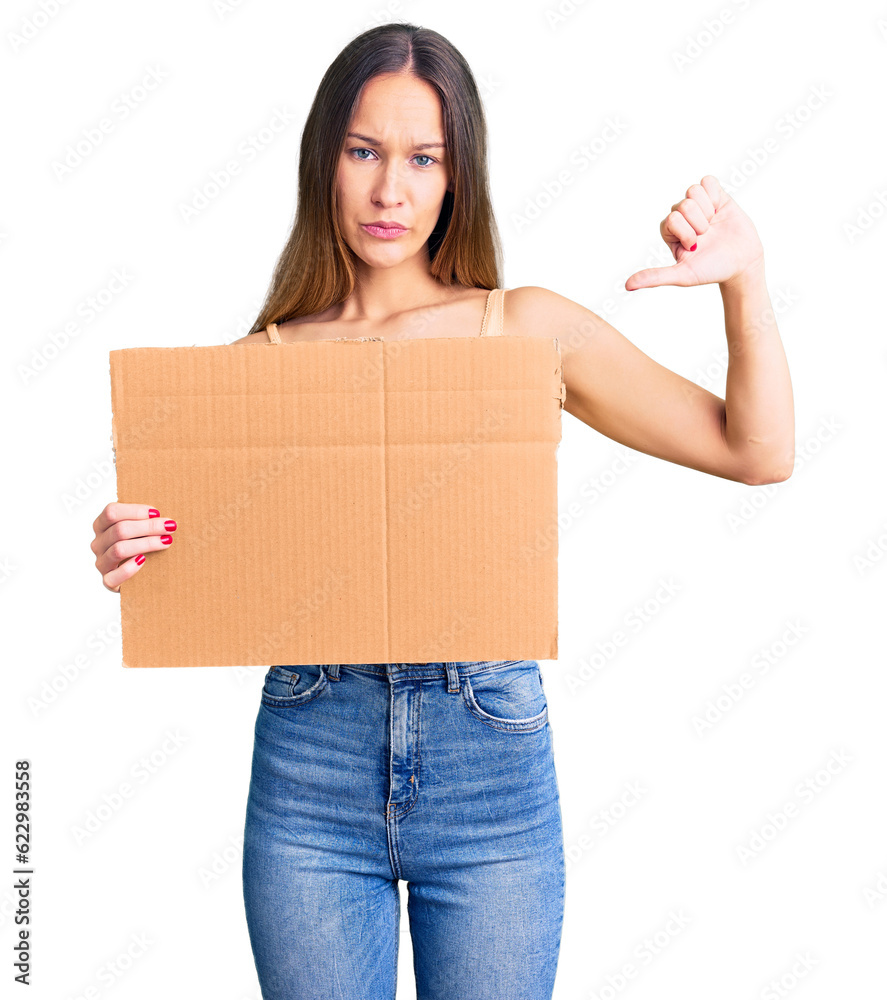 Beautiful brunette young woman holding cardboard banner with blank space with angry face, negative sign showing dislike with thumbs down, rejection concept Beautiful brunette young woman holding cardboard banner with blank space with angry face, negative sign showing dislike with thumbs down, rejection concept