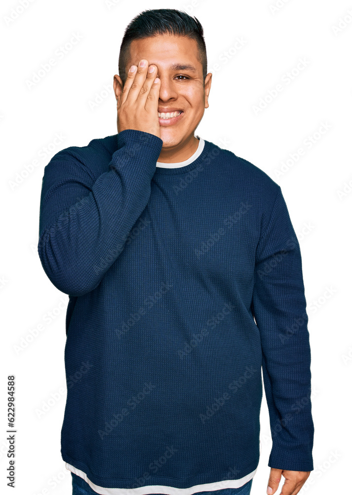 Young latin man wearing casual clothes covering one eye with hand, confident smile on face and surprise emotion. Young latin man wearing casual clothes covering one eye with hand, confident smile on face and surprise emotion.