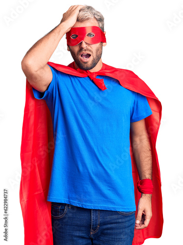 Young blond man wearing super hero custome surprised with hand on head for mistake, remember error. forgot, bad memory concept.