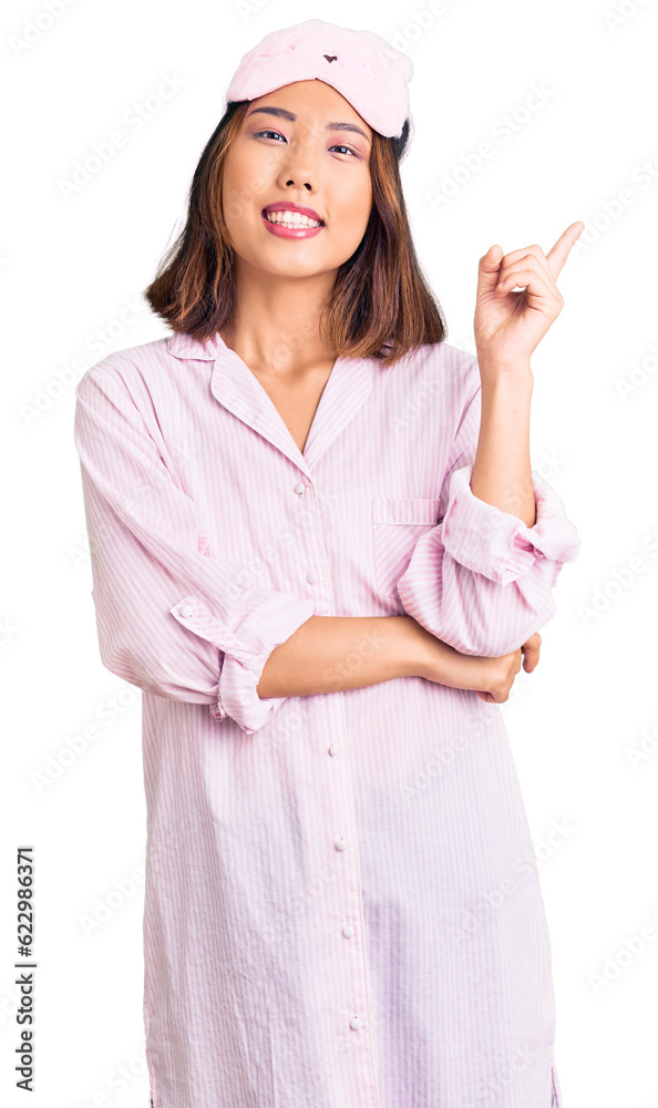 Young beautiful chinese girl wearing sleep mask and pajama with a big smile on face, pointing with hand and finger to the side looking at the camera.
