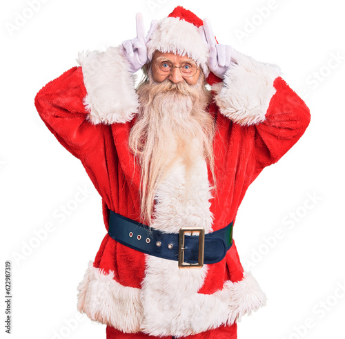 Old senior man with grey hair and long beard wearing traditional santa claus costume posing funny and crazy with fingers on head as bunny ears, smiling cheerful