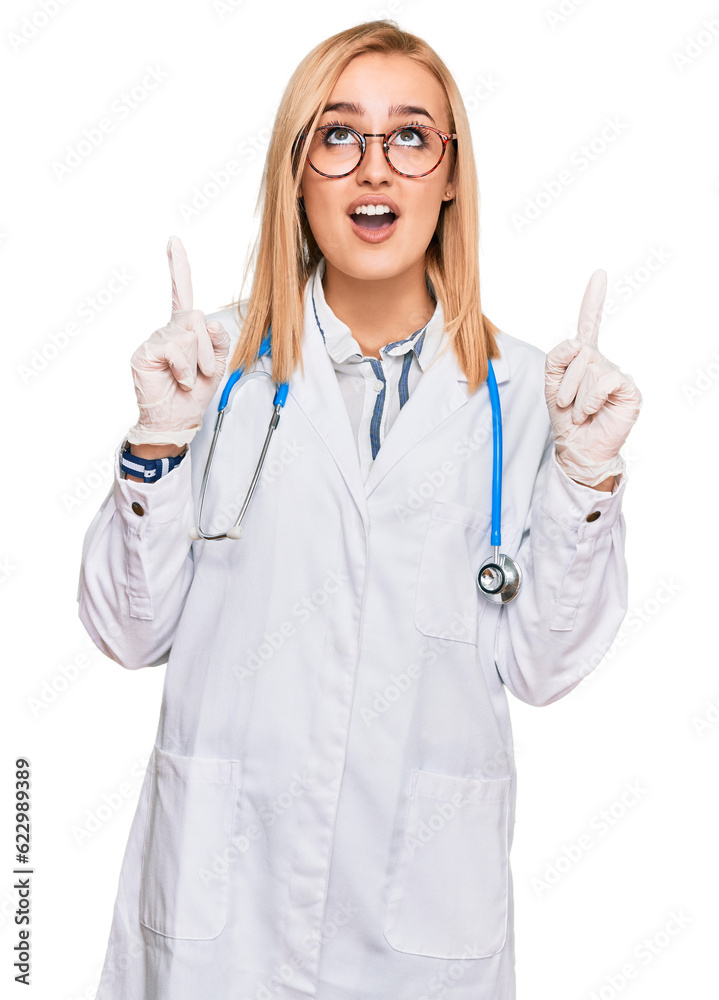 Beautiful caucasian woman wearing doctor uniform and stethoscope amazed and surprised looking up and pointing with fingers and raised arms. Beautiful caucasian woman wearing doctor uniform and stethoscope amazed and surprised looking up and pointing with fingers and raised arms.