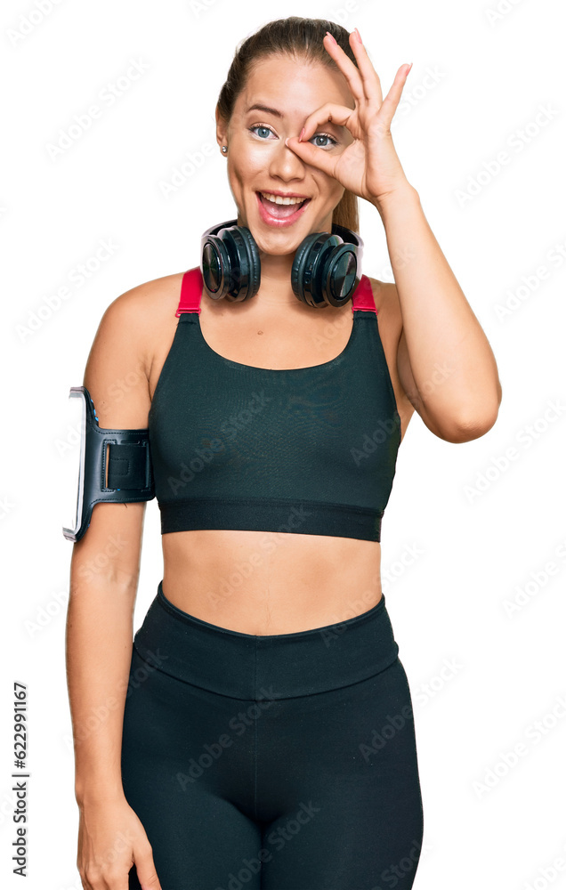Beautiful blonde woman wearing gym clothes and using headphones doing ok gesture with hand smiling, eye looking through fingers with happy face. Beautiful blonde woman wearing gym clothes and using headphones doing ok gesture with hand smiling, eye looking through fingers with happy face.