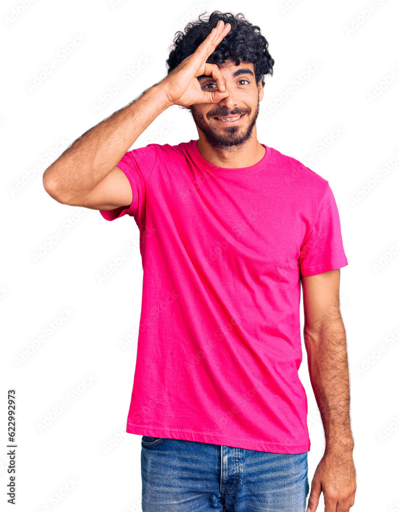 Handsome young man with curly hair and bear wearing casual pink tshirt doing ok gesture with hand smiling, eye looking through fingers with happy face. Handsome young man with curly hair and bear wearing casual pink tshirt doing ok gesture with hand smiling, eye looking through fingers with happy face.