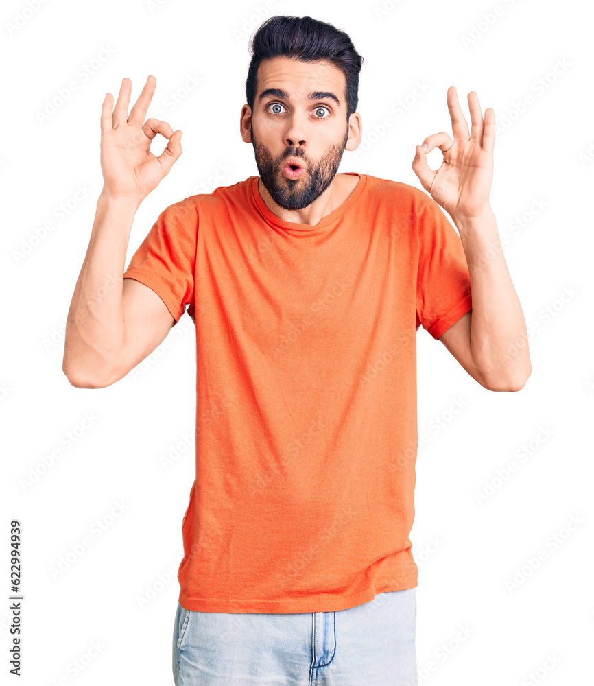 Young handsome man with beard wearing casual t-shirt looking surprised and shocked doing ok approval symbol with fingers. crazy expression Young handsome man with beard wearing casual t-shirt looking surprised and shocked doing ok approval symbol with fingers. crazy expression