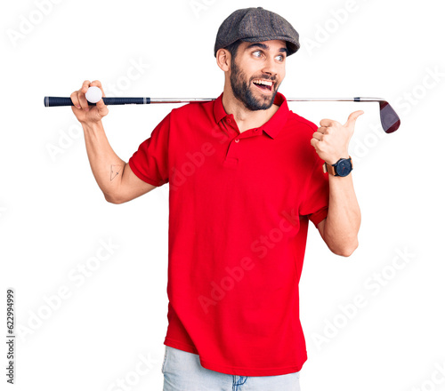 Young handsome man with beard playing golf holding club and ball pointing thumb up to the side smiling happy with open mouth