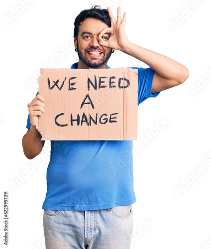 Young hispanic man holding we need a change banner smiling happy doing ok sign with hand on eye looking through fingers
