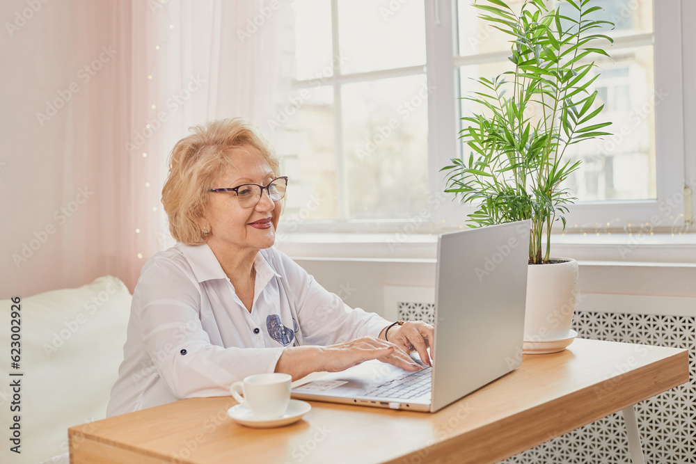 Portrait of charming cheerful person sit behind desktop with cup of coffee look interested tv, working from home indoors. Happy mature woman reading good news looking at laptop, watching online Portrait of charming cheerful person sit behind desktop with cup of coffee look interested tv, working from home indoors. Happy mature woman reading good news looking at laptop, watching online