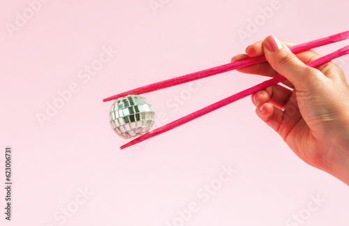 Female hand holding chopsticks and disco ball on pastel pink background. Retro fashion aesthetic party concept.