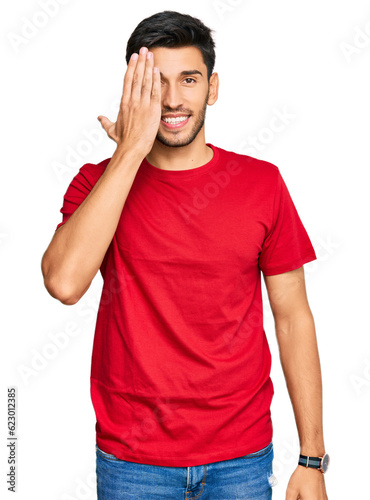 Young handsome man wearing casual red tshirt covering one eye with hand, confident smile on face and surprise emotion. © Krakenimages.com