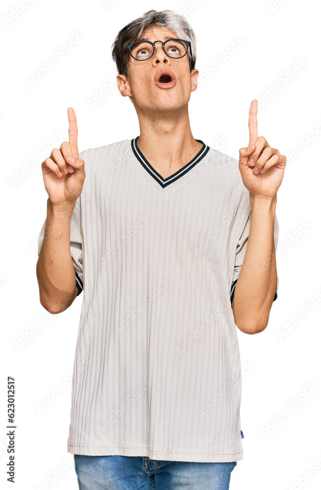 Young hispanic man wearing casual clothes and glasses amazed and surprised looking up and pointing with fingers and raised arms. Young hispanic man wearing casual clothes and glasses amazed and surprised looking up and pointing with fingers and raised arms.