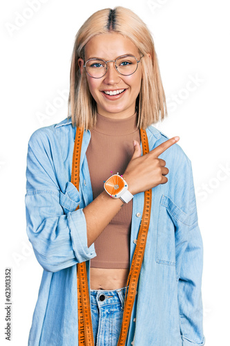 Beautiful young blonde woman dressmaker designer smiling cheerful pointing with hand and finger up to the side