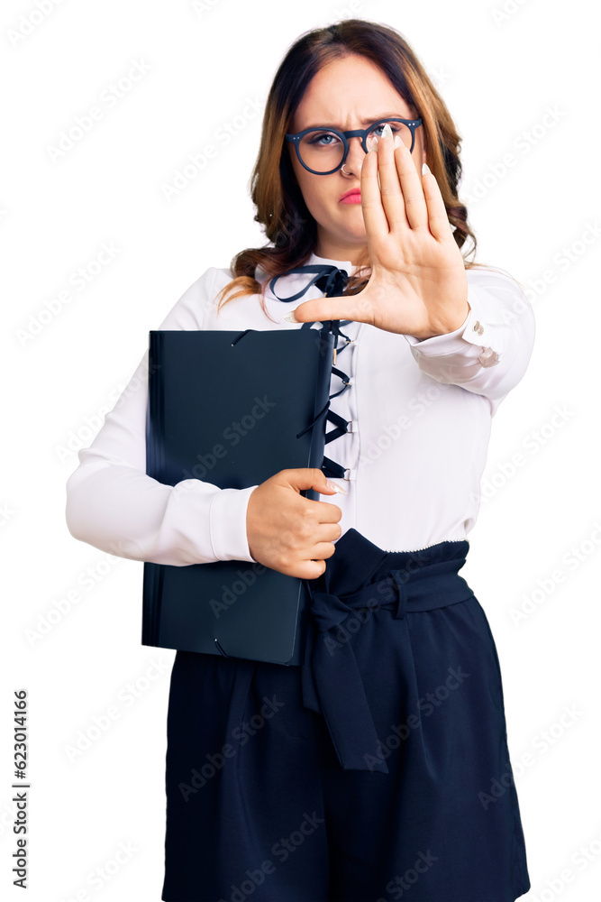 Young beautiful caucasian woman wearing business shirt and glasses holding folder with open hand doing stop sign with serious and confident expression, defense gesture Young beautiful caucasian woman wearing business shirt and glasses holding folder with open hand doing stop sign with serious and confident expression, defense gesture