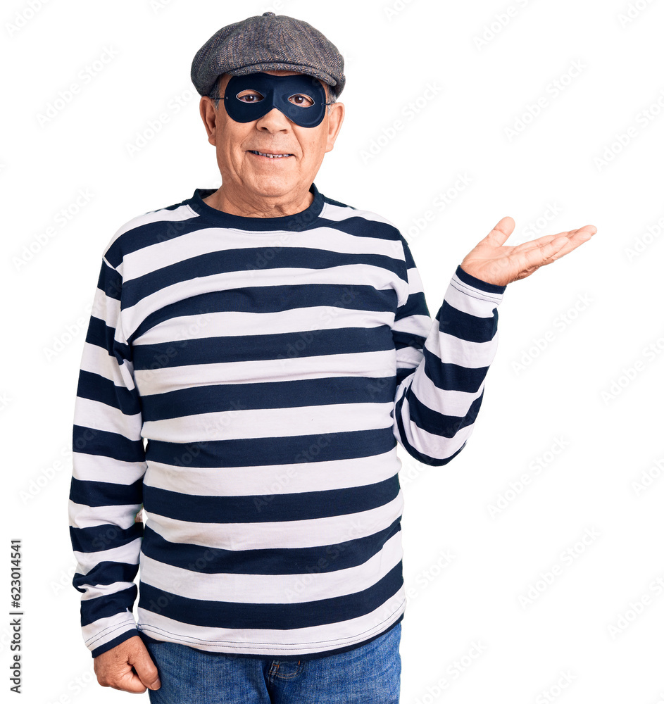 Senior handsome man wearing burglar mask and t-shirt smiling cheerful presenting and pointing with palm of hand looking at the camera. Senior handsome man wearing burglar mask and t-shirt smiling cheerful presenting and pointing with palm of hand looking at the camera.