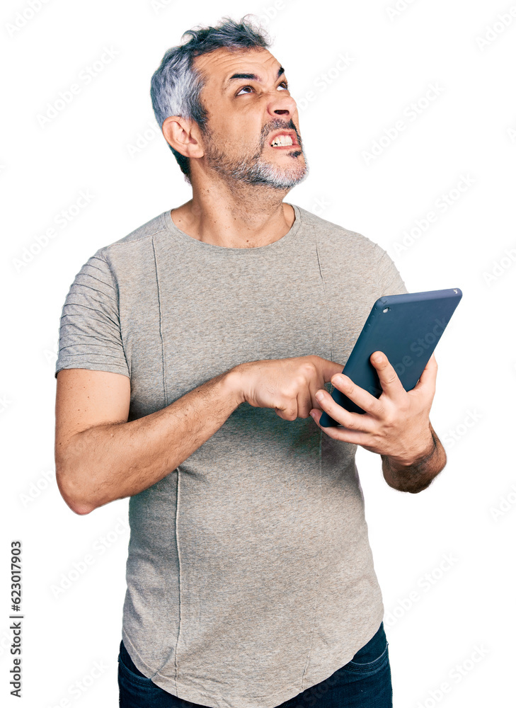 Middle age hispanic with grey hair using touchpad device angry and mad screaming frustrated and furious, shouting with anger looking up. Middle age hispanic with grey hair using touchpad device angry and mad screaming frustrated and furious, shouting with anger looking up.