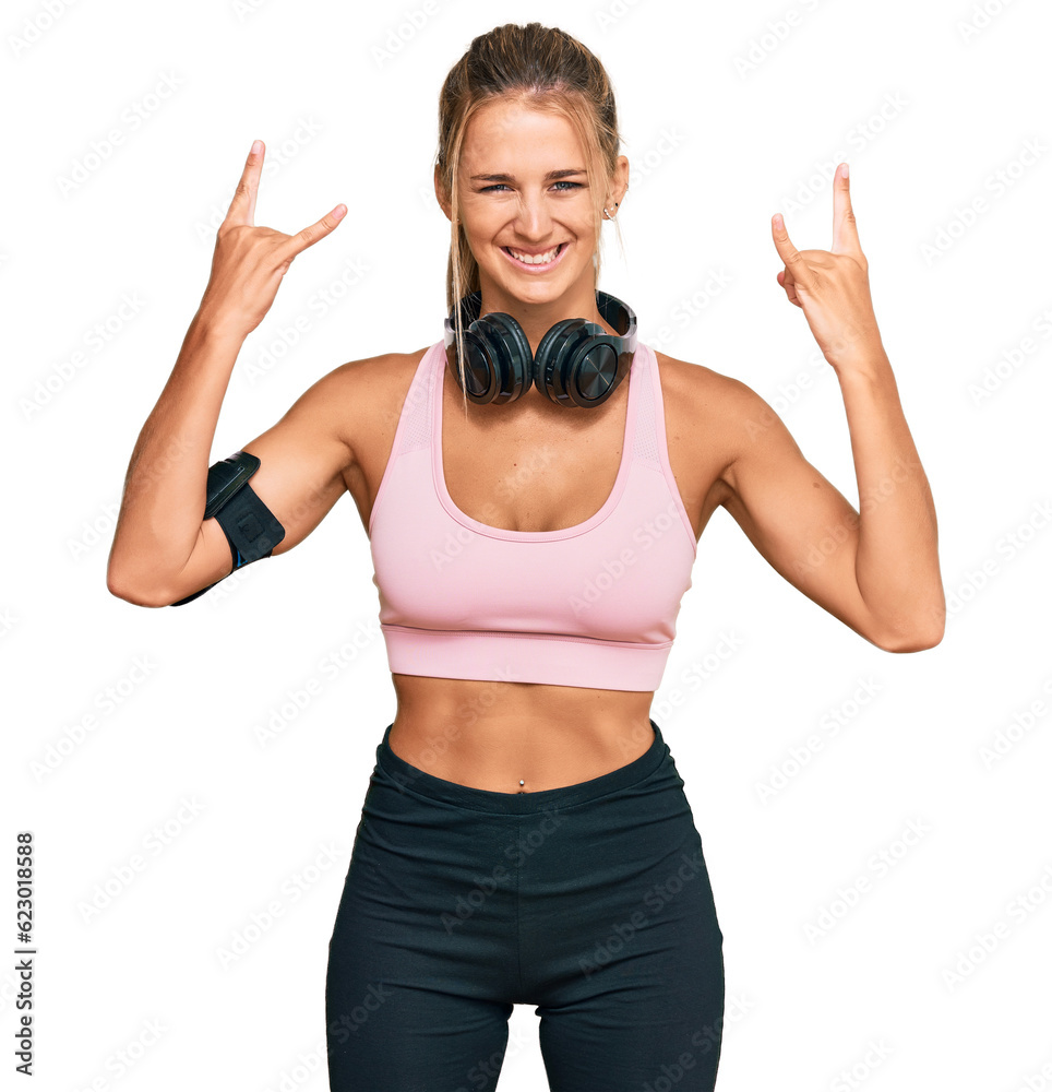 Young blonde woman wearing gym clothes and using headphones shouting with crazy expression doing rock symbol with hands up. music star. heavy concept. Young blonde woman wearing gym clothes and using headphones shouting with crazy expression doing rock symbol with hands up. music star. heavy concept.