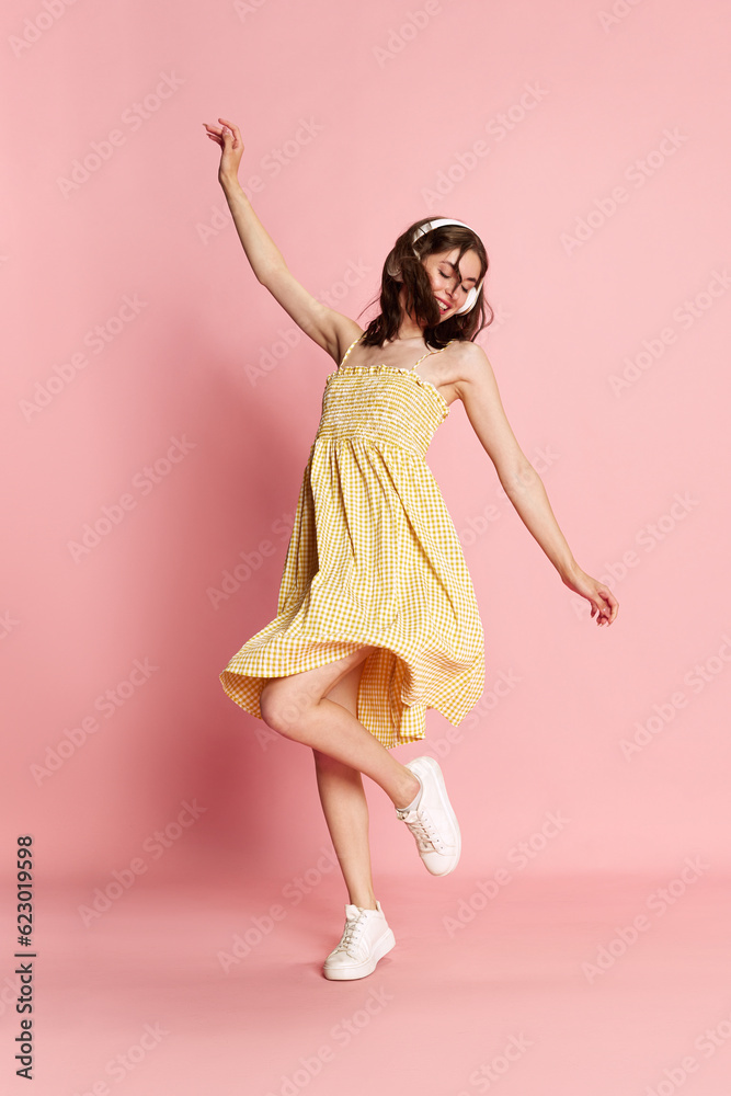 Full-length image of young pretty girl in yellow dress listening to music in headphones and dancing over pink studio background. Concept of human emotions, fashion, beauty, lifestyle, summer vibe, ad