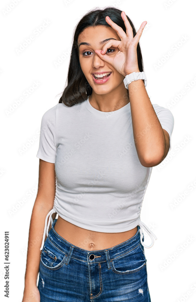 Young hispanic girl wearing casual white t shirt doing ok gesture with hand smiling, eye looking through fingers with happy face.