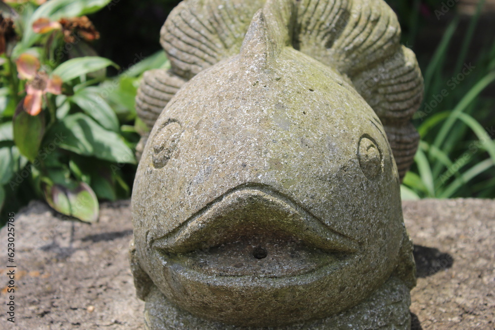 A photo of a stone statue of a fish against a background of green plants and a muddy floor. 