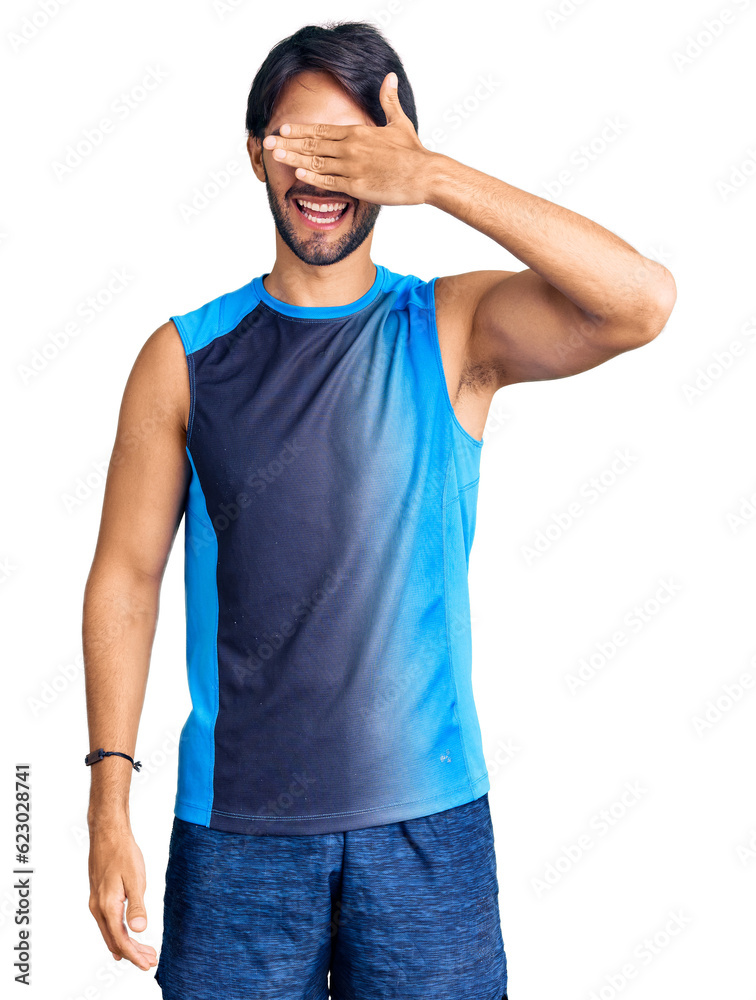 Handsome hispanic man wearing sportswear smiling and laughing with hand on face covering eyes for surprise. blind concept. Handsome hispanic man wearing sportswear smiling and laughing with hand on face covering eyes for surprise. blind concept.